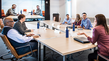 a group of coworkers in a meeting room video-chatting with colleagues from a different location on the office TV screen